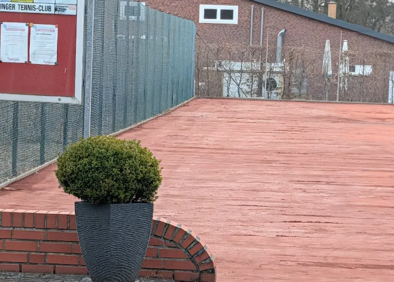 Rote Sandfläche neben dem Eingang, vorne ein Buchsbaum im Kübel auf einer Backsteinmauer.