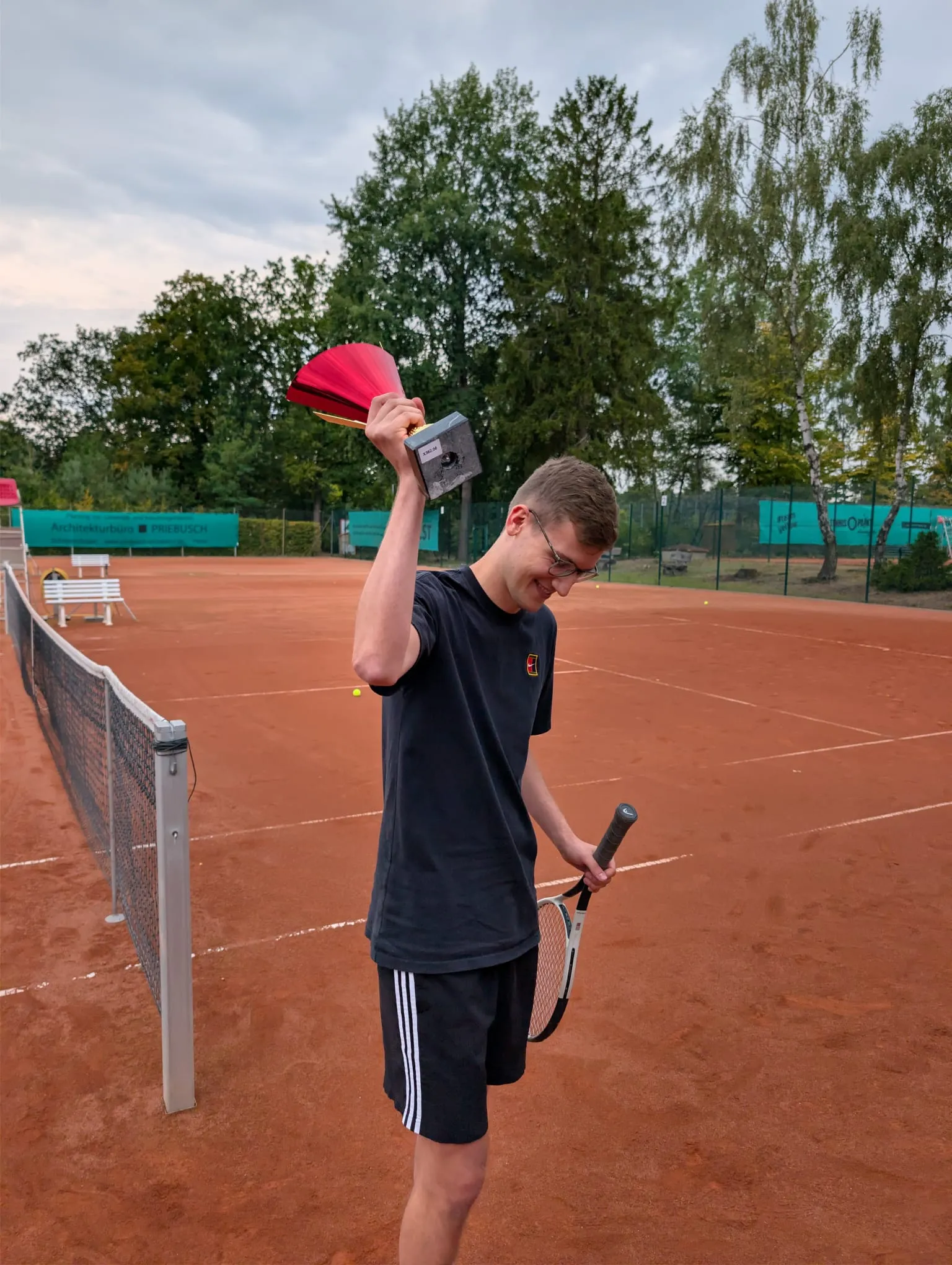 Tom Eisele hält auf dem Sandplatz lächelnd den rot-goldenen Pokal hoch, in der anderen Hand den Schläger.