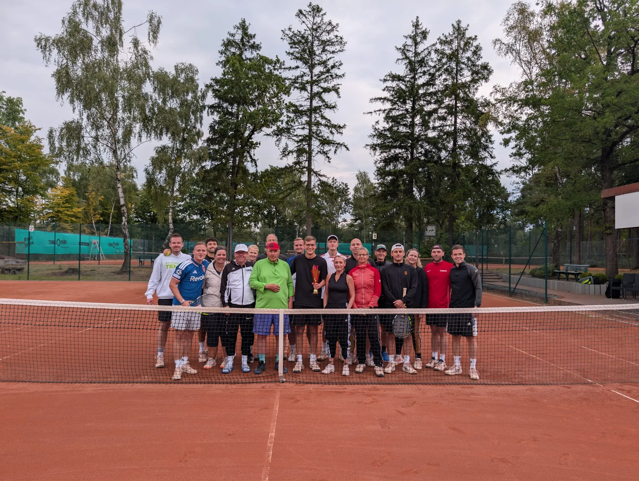 Gruppenfoto der Teilnehmenden hinter dem Netz auf dem Sandplatz, in der Mitte Tom Eisele mit dem Pokal.