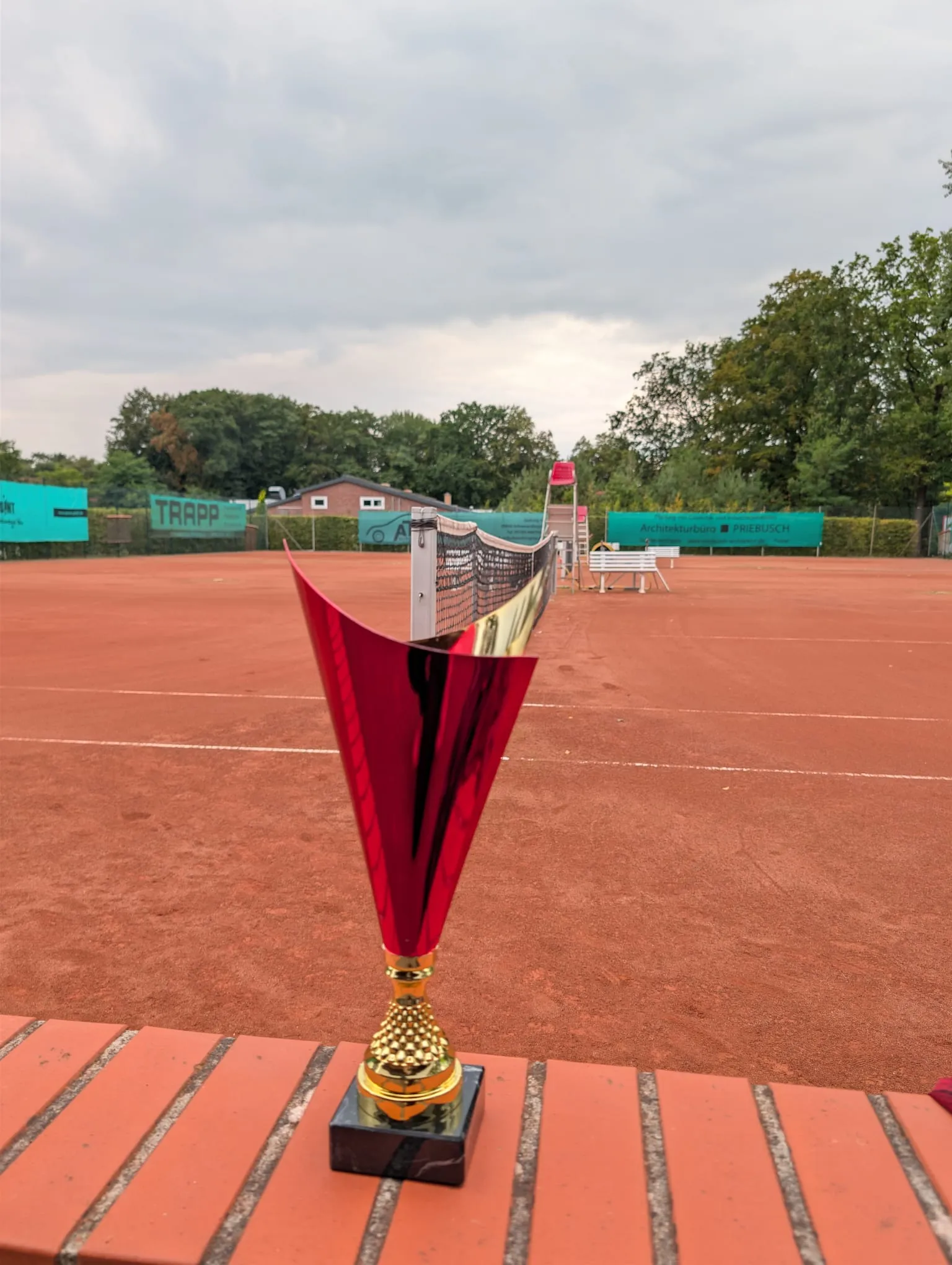 Rot-goldener Pokal auf einer Backsteinmauer, dahinter Sandplatz und Netz unter grauem Himmel.